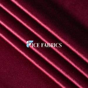 Puede incluir: Una tela de terciopelo color burdeos intenso con una textura suave que refleja la luz y pliegues diagonales. La tela está superpuesta con un logotipo blanco que dice ICE FABRICS, junto a un icono gráfico circular en azul claro y blanco.