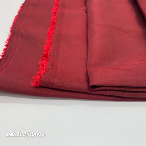 May include: Close-up of a folded piece of burgundy fabric with a red fringe trim. The fabric has a subtle textured pattern.