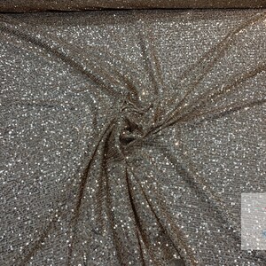 May include: A close-up of a silver sequined fabric with a brown undertone. The sequins are small and densely packed, creating a shimmering effect.