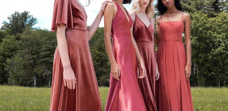 May include: Four women wearing different styles of long, rust-colored bridesmaid dresses. The dresses are all floor-length and have a variety of necklines and straps.