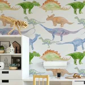 May include: A watercolor dinosaur wallpaper pattern with a variety of dinosaurs in shades of green, blue, orange, and purple. The wallpaper is shown in a room with a white shelf and a wooden desk.
