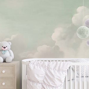 May include: A white cot with a white and grey patterned blanket. A grey teddy bear sits on a wooden chest of drawers with the word "LOVE" spelled out in wooden blocks. The wall behind the cot is painted with a light green and white cloudy sky design with hanging felt balls in shades of green and purple.