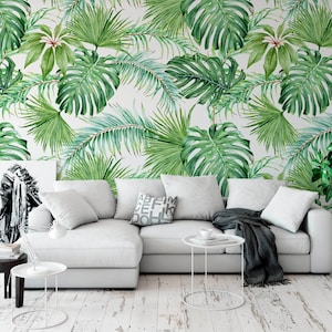 May include: A white sectional sofa with a gray throw blanket and a white side table with a cup on it. The sofa is in front of a wall with a tropical leaf wallpaper pattern.