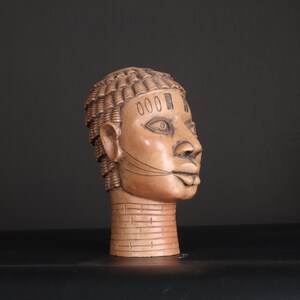 Puede incluir: Una escultura de madera de una cabeza humana con un peinado y rasgos faciales detallados. La escultura está pintada en tonos marrones y negros.