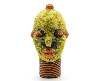 May include: A beaded sculpture of a human head with a yellow face, orange lips, black eyes, and a black nose. The head is decorated with orange and black beads around the neck.