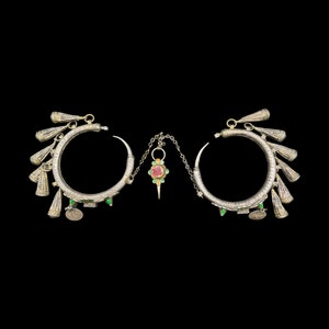 May include: A pair of silver-toned hoop earrings with intricate detailing and dangling charms. The earrings feature a central pendant with a red and green floral design, connected by a delicate chain. The design includes small green accents and coin-like charms.