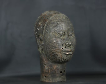 May include: A bronze sculpture of a human head with a detailed, textured surface. The head is missing the lower jaw and has several holes throughout. The sculpture is dark brown in color and has a patina finish.