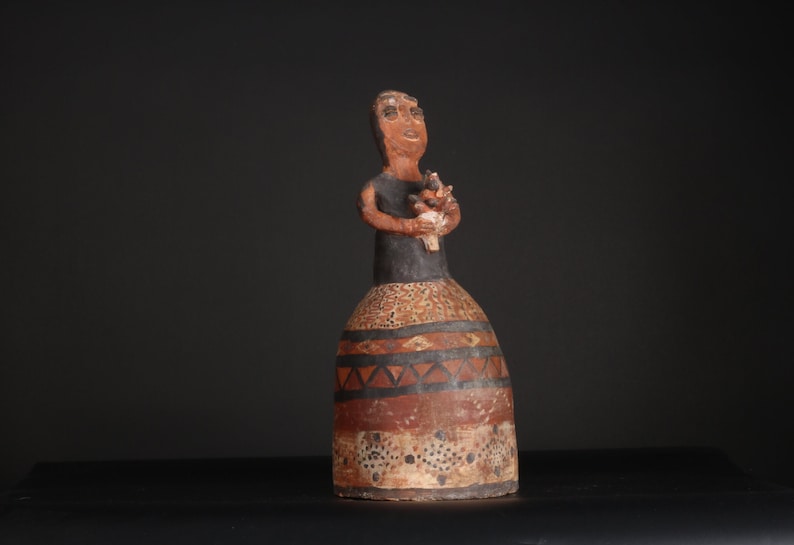 May include: A ceramic figurine of a woman holding a small figure in her arms. The figurine is painted in black, brown, and white with geometric patterns.