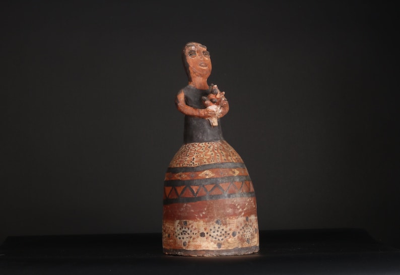 May include: A terracotta figurine of a woman holding a bouquet of flowers. The figurine is painted in black, brown, and white with geometric patterns.
