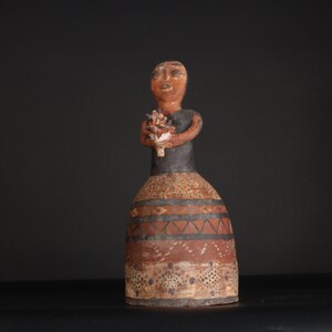 May include: A ceramic figurine of a woman holding a bouquet of flowers. The figurine is painted in brown, black, and white with geometric patterns on the skirt.