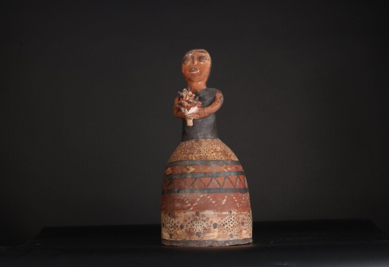 May include: A ceramic figurine of a woman holding a bouquet of flowers. The figurine is painted in brown, black, and white with geometric patterns on the skirt.