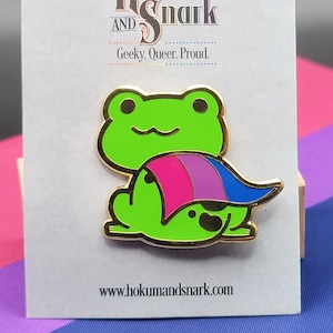 Puede incluir: Una rana verde con una capa de bandera bi. La rana está sentada en una tarjeta blanca con el texto "Hokum and Snark, Geeky, Queer, Proud. www.hokumandsnark.com"