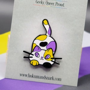 May include: A yellow, purple, and black enamel pin of a cat with a white face and yellow eyes. The cat is curled up and looking to the left. The pin is on a white card with the text "Hokum and Snark" in black and the text "Geeky. Queer. Proud." in black. The card also has the website "www.hokumandsnark.com" in black.