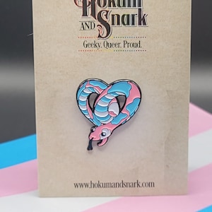 Op de afbeelding: Een roze en blauwe emaille pin met een slang die in de vorm van een hart is opgerold. De pin zit op een kaart met de tekst "Hokum and Snark, Geeky. Queer. Proud." en "www.hokumandsnark.com".