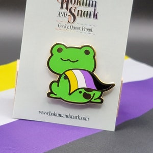 Pode incluir: Um sapo verde vestindo uma capa com as cores da bandeira do orgulho não binário. O sapo está em um cartão branco com o texto "Hokum and Snark, Geeky, Queer, Proud" e o endereço do site "www.hokumandsnark.com". O sapo está sentado em um tecido listrado roxo, branco, amarelo e preto.