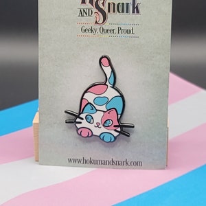 May include: A pink, blue and white enamel pin of a cat with a transgender pride flag in the background. The pin says "Hokum and Snark" and "Geeky. Queer. Proud." at the top and "www.hokumandsnark.com" at the bottom.