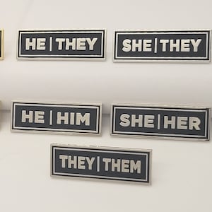 Puede incluir: Un conjunto de ocho pines de esmalte con texto negro sobre un fondo dorado o plateado. Los pines presentan diferentes combinaciones de pronombres, incluyendo "SHE | THEY", "HE | THEY", "SHE | HER", "HE | HIM" y "THEY | THEM".