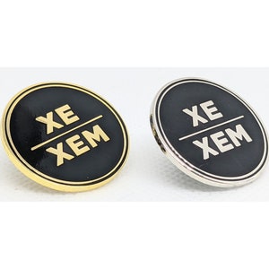 Könnte beinhalten: Zwei runde Pins mit schwarzem Hintergrund und goldener Beschriftung. Die Pins zeigen "XE/XEM". Ein Pin ist golden und der andere ist silber.