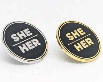 Può includere: Due spille rotonde con sfondo nero e lettere bianche. Il testo sulle spille recita "SHE HER". Una spilla ha una finitura argentata e l'altra una finitura dorata.