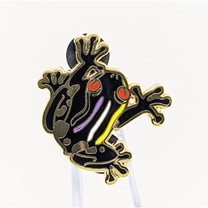 May include: A black frog pin with gold trim and accents. The frog has red eyes, and a white, purple, and yellow stripe design. The pin is on a clear plastic stand against a white background.