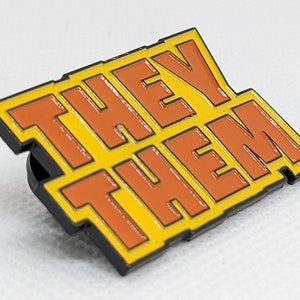 Puede incluir: Un pin de esmalte amarillo y naranja con las palabras "THEY THEM" en letras negras delineadas.