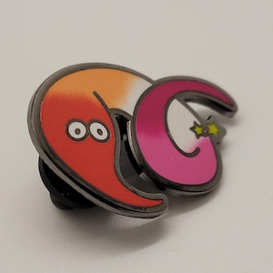 May include: A red and orange worm with two eyes and a pink and white worm with a yellow star on its tail. The worms are attached to a black metal pin back.