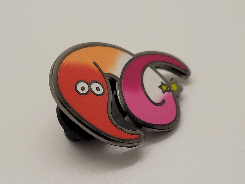 May include: A red and orange worm with two eyes and a pink and white worm with a yellow star on its tail. The worms are attached to a black metal pin back.