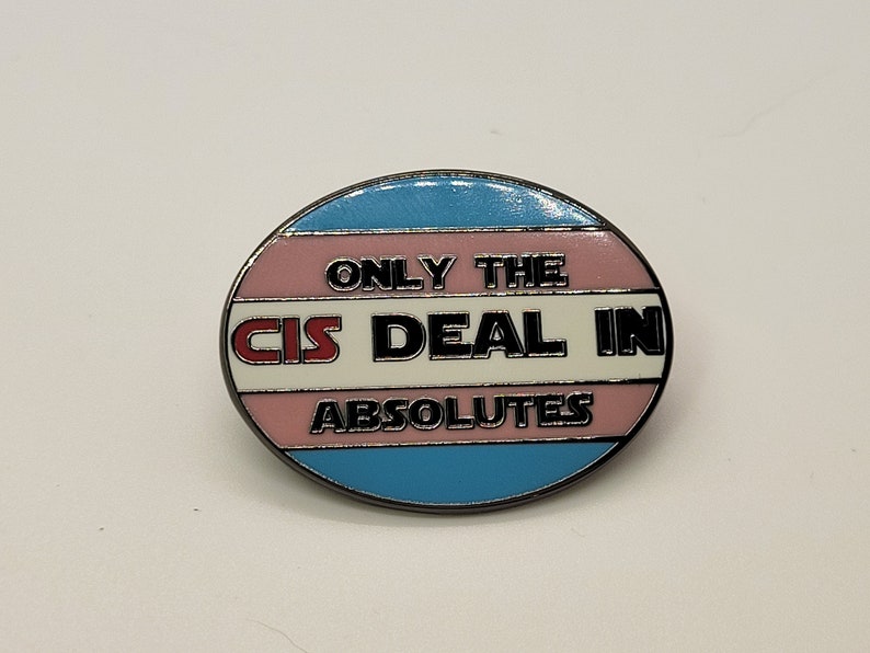 May include: A round pin with a transgender pride flag design. The pin says "Only the cis deal in absolutes" in black text on a white background.