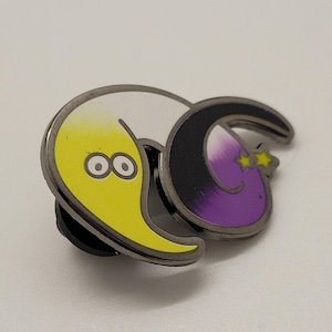 May include: A yellow and purple crescent moon pin with a black outline. The yellow crescent moon has two eyes and a smile. The purple crescent moon has two stars above it and a smile.