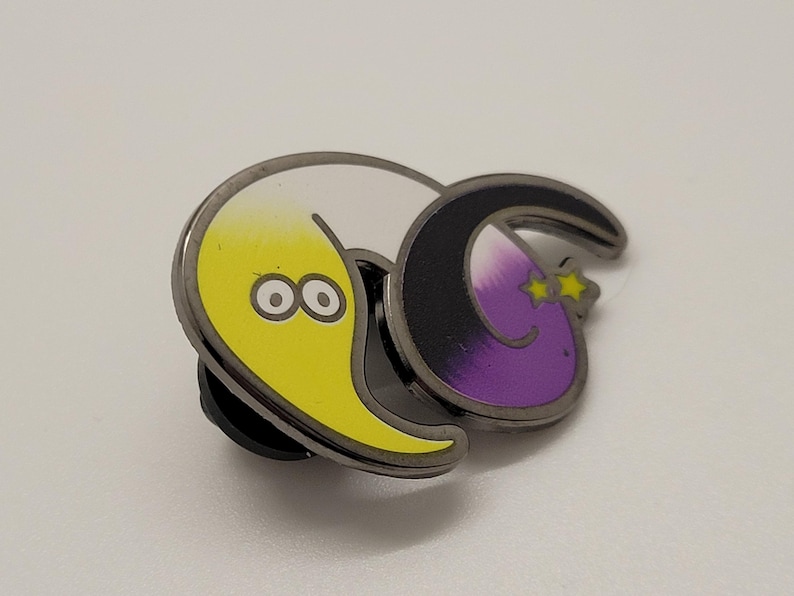 May include: A yellow and purple crescent moon pin with a black outline. The yellow crescent moon has two eyes and a smile. The purple crescent moon has two stars above it and a smile.