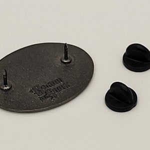 May include: The back of a silver oval-shaped pin with the text "WOMAN IN THE STINK" in a gothic font. The pin has two black rubber pin backs.