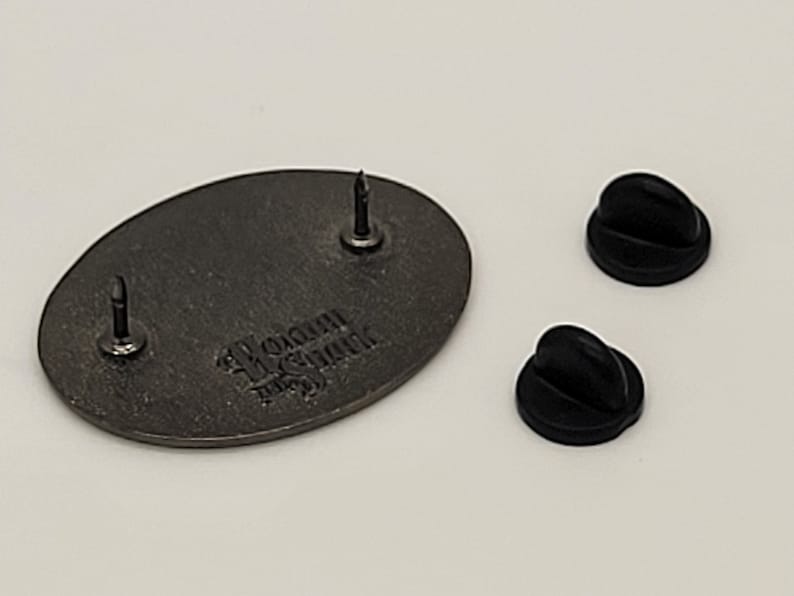 May include: The back of a silver oval-shaped pin with the text "WOMAN IN THE STINK" in a gothic font. The pin has two black rubber pin backs.