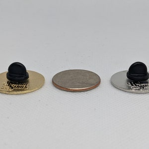 K&ouml;nnte beinhalten: Drei Emaille-Pins mit schwarzen Gummib&ouml;den. Die Pins haben das gleiche Design, aber in verschiedenen Ausf&uuml;hrungen: Gold, Silber und Nickel. Die Pins sind etwa so gro&szlig; wie eine US-amerikanische 25-Cent-M&uuml;nze.