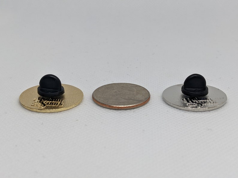 K&ouml;nnte beinhalten: Drei Emaille-Pins mit schwarzen Gummib&ouml;den. Die Pins haben das gleiche Design, aber in verschiedenen Ausf&uuml;hrungen: Gold, Silber und Nickel. Die Pins sind etwa so gro&szlig; wie eine US-amerikanische 25-Cent-M&uuml;nze.