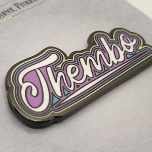 May include: A purple, blue, yellow, and white enamel pin with the word "Thembe" in a retro, stylized font.