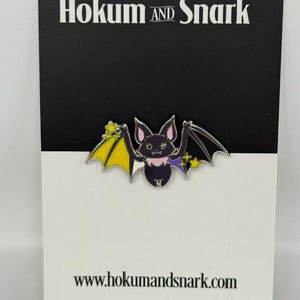 May include: A metal enamel pin shaped like a bat with wings colored in yellow, white, black, and purple, featuring small yellow stars. The pin is attached to a black and white display card with the text Hokum AND Snark and www.hokumandsnark.com.