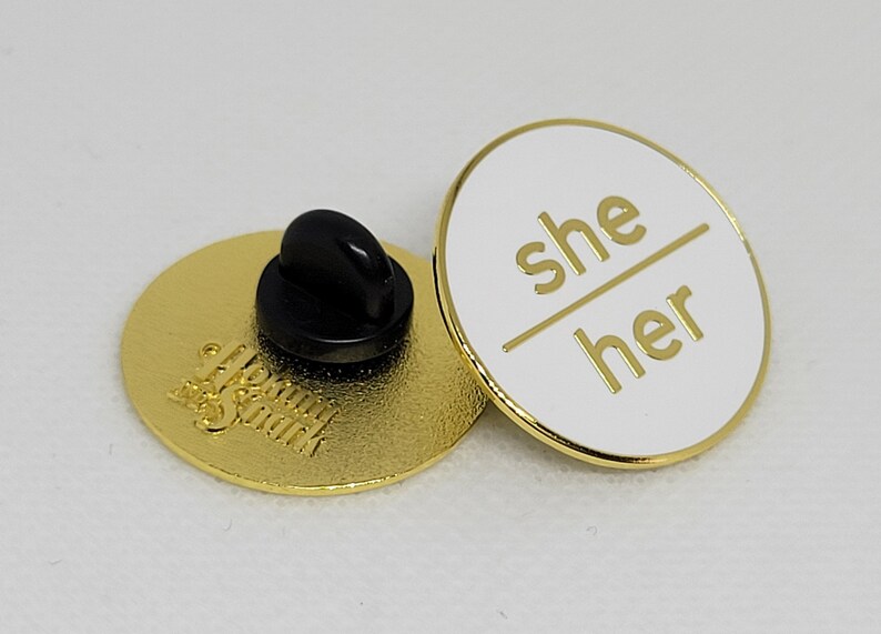 K&ouml;nnte beinhalten: Eine wei&szlig;e Emaille-Anstecknadel mit goldener Beschriftung, die "she/her" lautet. Die R&uuml;ckseite der Anstecknadel ist golden mit den Worten "Meow & Spark" in einer kursivschriftlichen Schrift.