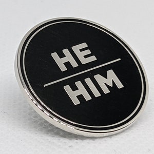 May include: A silver-colored pin with a black background and the words "HE HIM" in white lettering.
