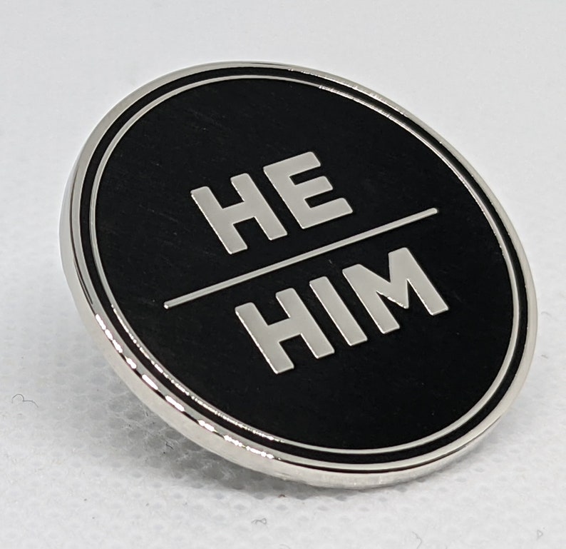 May include: A silver-colored pin with a black background and the words "HE HIM" in white lettering.