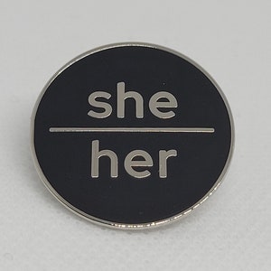 K&ouml;nnte beinhalten: Eine schwarze und silberne Anstecknadel mit den Worten "she/her" in wei&szlig;en Buchstaben.