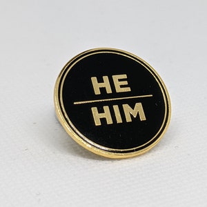 May include: A black and gold circular pin with the words "HE HIM" in gold lettering.