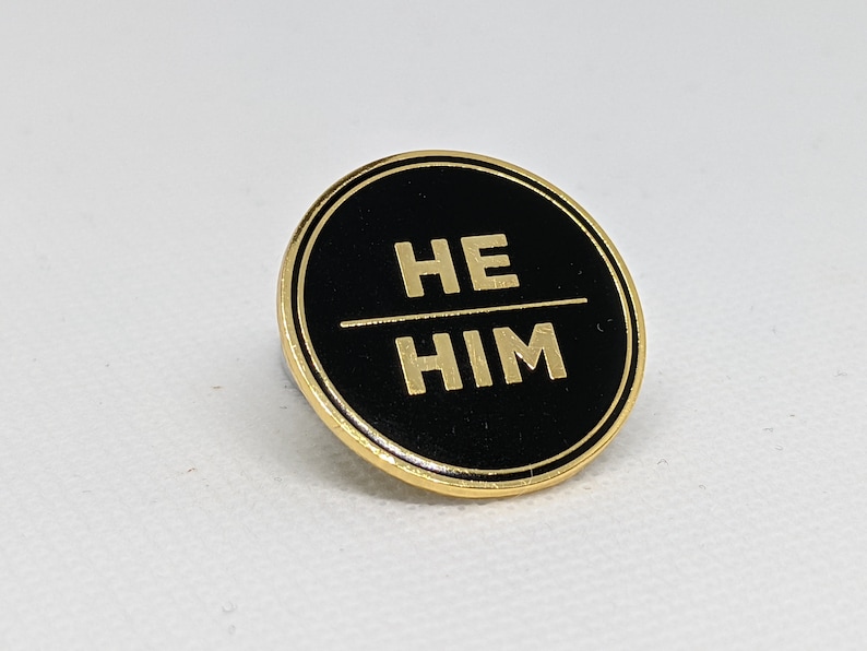 May include: A black and gold circular pin with the words "HE HIM" in gold lettering.