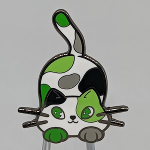 May include: A white, green, and black enamel pin of a cartoon cat with big eyes. The cat is sitting on its hind legs with its tail curled up behind it.
