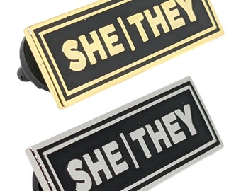 May include: Two enamel pins with black backgrounds and gold and silver borders. The pins say "SHE|THEY" in white and gold lettering.