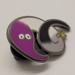May include: A black and white worm with a purple worm with two eyes on top. The worm is curled up in a circle. The worm is a metal pin.