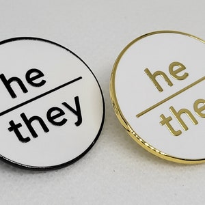 May include: Two white enamel pins with black and gold outlines. The pins are round and feature the text "he/they" in black and gold lettering.