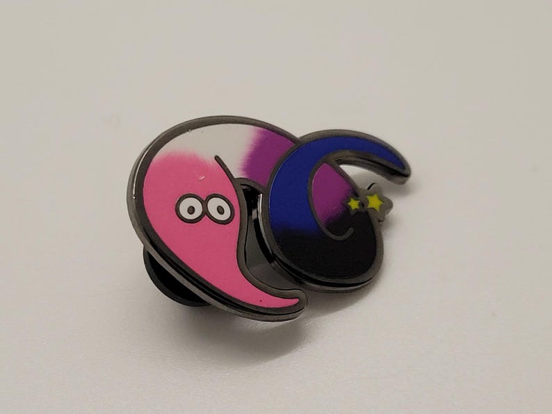 May include: A pink and purple worm with googly eyes is curled around a blue and black crescent moon with yellow stars. The worm and moon are attached to a metal pin back.