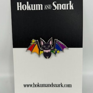 May include: A black enamel bat pin with rainbow-colored wings, featuring small yellow stars on the wing tips. The pin is attached to a white and black display card with the text Hokum AND Snark at the top and www.hokumandsnark.com at the bottom.