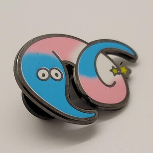 May include: A blue and pink worm with a white stripe and a yellow star on its tail. The worm has two large black eyes and is curled up in a circle. The worm is a metal pin.