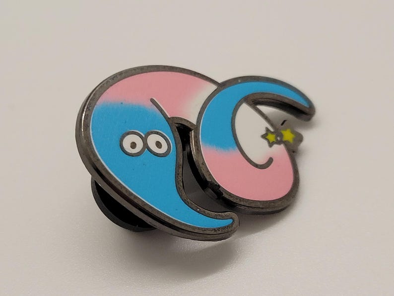 May include: A blue and pink worm with a white stripe and a yellow star on its tail. The worm has two large black eyes and is curled up in a circle. The worm is a metal pin.